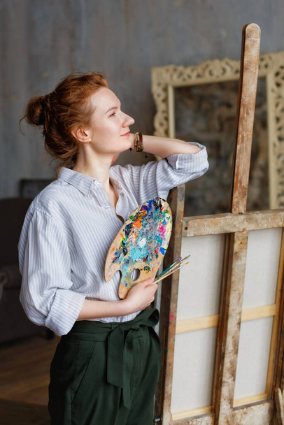Portret van een roodharige professionele kunstenaar met penseel en palet, op doek geschreven in de studio. Een vrouwelijke kunstenaar tekent op de werkplek. - Foto, afbeelding