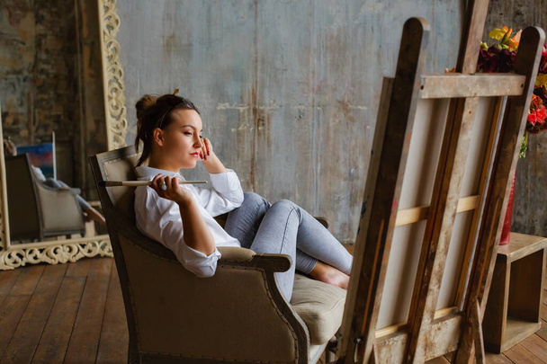 Retrato de un artista profesional pintando sobre lienzo en un estudio en casa. Una artista femenina dibuja. Una chica con gafas, una camisa blanca y jeans con pincel y paleta. - Foto, Imagen