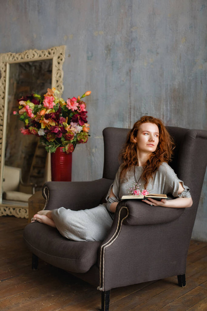 Portret van een jonge aantrekkelijke roodharige vrouw die een boek leest in een knusse grijze stoel. Er is een vaas bloemen in de buurt.. - Foto, afbeelding