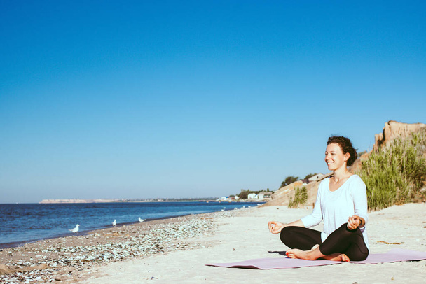 Yoga en la playa. Mujer joven sonriente sentada en pose de loto, practicando técnicas de respiración cerca del océano sobre fondo azul del cielo. Meditación y pranayama. Estilo de vida saludable de verano. Copiar espacio. - Foto, Imagen