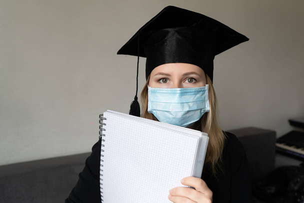 Студент в маске одет в выпускной плащ и халат, держащий в руках пустое место для копирования концепции коронавирусной пандемии ковида класса 2021. Выпускник на белом фоне - Фото, изображение