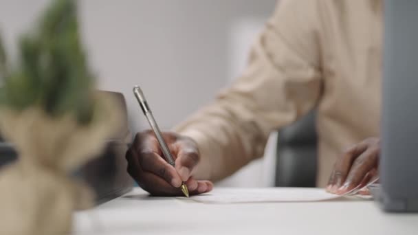 бизнесмен делает заметки, пишет на бумаге, сидит за столом в рабочей комнате, крупным планом смотрит на руку афро-американца - Кадры, видео