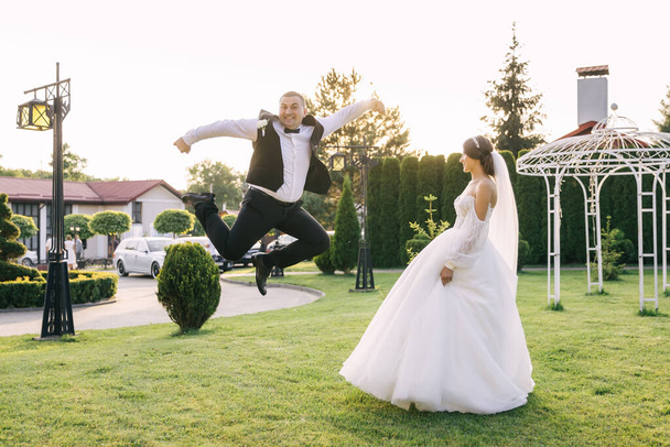 Счастливые улыбающиеся стильные невеста и жених прогуливаются в летнем зеленом парке, мужчина в прыжке, веселясь в день своей свадьбы. - Фото, изображение
