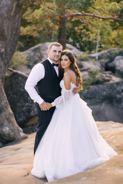 jovem casal bonito de homem e mulher em vestido de noiva branco e terno andando abraçando enquanto está em pé nas montanhas de pedra. - Foto, Imagem