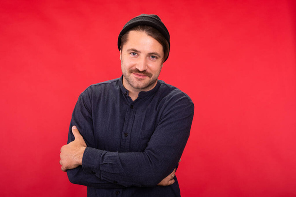 Smiling Handsome unshaven guy in knitted cap posing crossed arms on red studio - Photo, Image