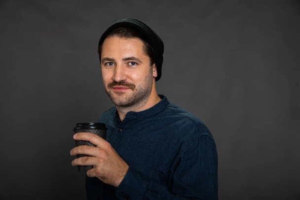Glimlachende ongeschoren man poseren met wegwerp koffie kopje op grijze studio achtergrond - Foto, afbeelding