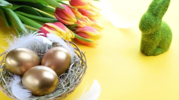 Huevos de Pascua oro. Huevo dorado en cesta con tulipanes primaverales, plumas blancas sobre fondo amarillo pastel en feliz decoración de Pascua. Diseño de Pascua de felicitación - Metraje, vídeo