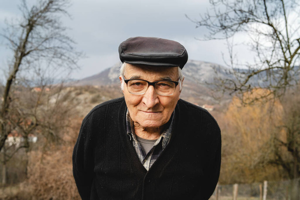 Baharda ya da sonbaharda ayakta duran yaşlı beyaz adamın portresi. Çiftliğin kırsalında dikilip kameraya bakan neşeli erkek çiftçinin portresi. Gerçek insanlar kavramı. - Fotoğraf, Görsel