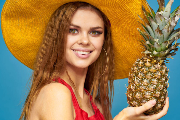 Sarı şapkalı, ananas elli, kıvırcık saçlı egzotik meyveli, mavi arka planlı çekici görünümlü bir kadın. - Fotoğraf, Görsel
