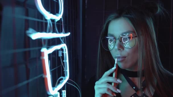 Confident and elegant Caucasian woman with glasses in purple blue light smokes an electronic cigarette at a party near neon lights. Produces a lot of smoke. Cute female close-up - Footage, Video