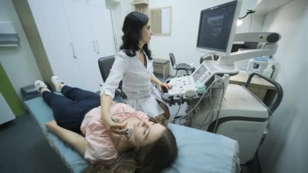 Diagnóstico por ultrasonido de la glándula tiroides joven en la clínica. Médico mujer pasa sensor de ultrasonido sobre el cuello del paciente. - Imágenes, Vídeo