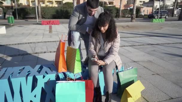 Young happy shopaholic multiracial couple having fun outdoor in town with internet on smartphone to check shopping online deals among many colored paper bags. New way to buy due to remote connection - Footage, Video