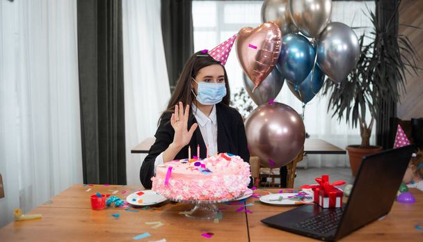 portrait d'une jeune belle femme caucasienne assise à une table et célébrant son anniversaire. Un masque médical est porté sur le visage. Communiquer via webcam et rire joyeusement. Il agite sa main en saluant. - Photo, image