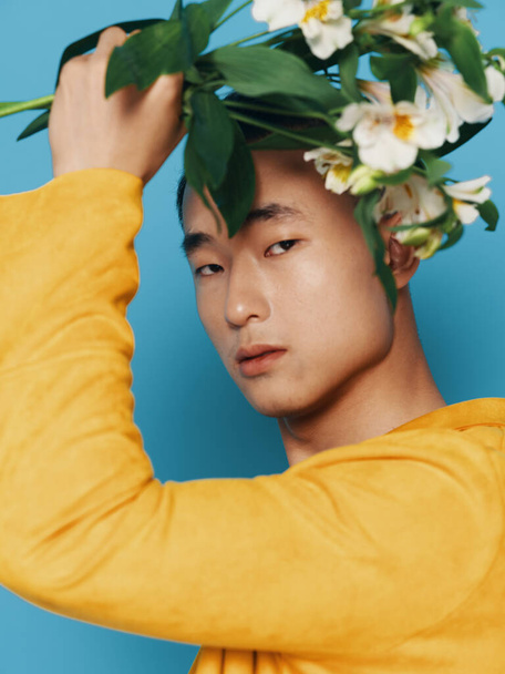 Encantador tipo con un ramo de flores blancas sobre su cabeza Sobre un fondo azul en un abrigo amarillo - Foto, Imagen