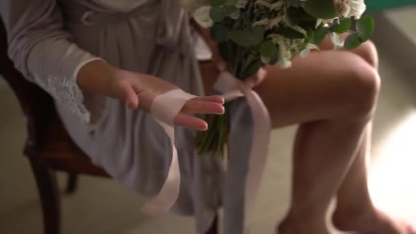 Een vrouw in een zijden badjas houdt een bruidsboeket op schoot en speelt met lange linten - Video