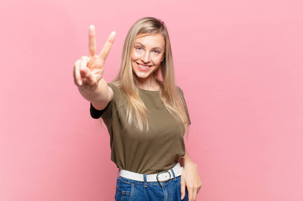 young blond pretty woman smiling and looking happy, carefree and positive, gesturing victory or peace with one hand - Photo, image