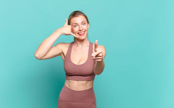 young blond woman smiling cheerfully and pointing to camera while making a call you later gesture, talking on phone. sport concept - Foto, afbeelding