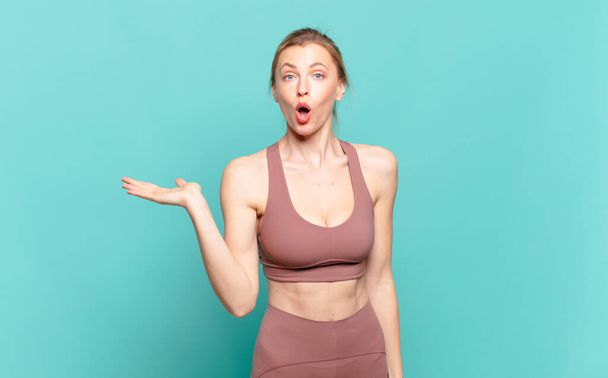 young blond woman looking surprised and shocked, with jaw dropped holding an object with an open hand on the side. sport concept - Фото, изображение