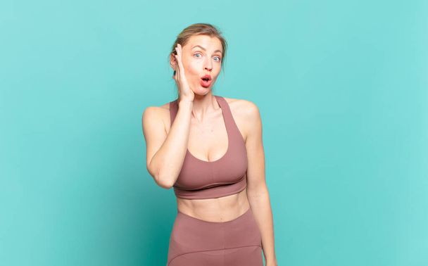 young blond woman feeling shocked and astonished holding face to hand in disbelief with mouth wide open. sport concept - Фото, изображение