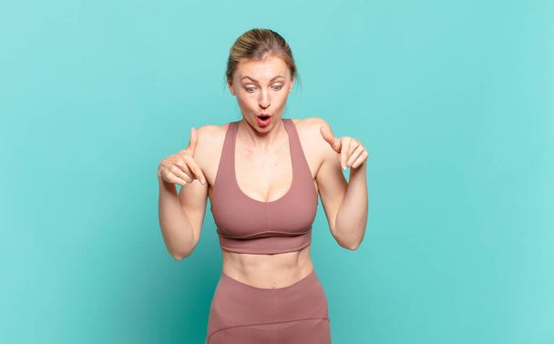 young blond woman feeling shocked, open-mouthed and amazed, looking and pointing downwards in disbelief and surprise. sport concept - Zdjęcie, obraz