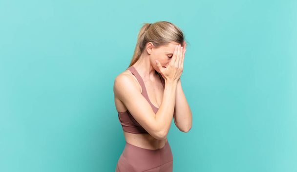 young blond woman covering eyes with hands with a sad, frustrated look of despair, crying, side view. sport concept - 写真・画像
