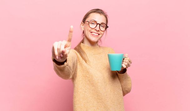 young blond woman smiling proudly and confidently making number one pose triumphantly, feeling like a leader. coffee concept - Foto, imagen