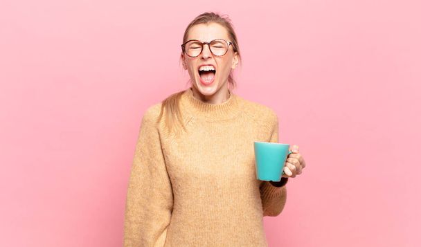 young blond woman shouting aggressively, looking very angry, frustrated, outraged or annoyed, screaming no. coffee concept - Photo, Image