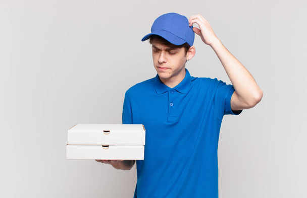 young pizza deliver boy feeling puzzled and confused, scratching head and looking to the side - Фото, изображение