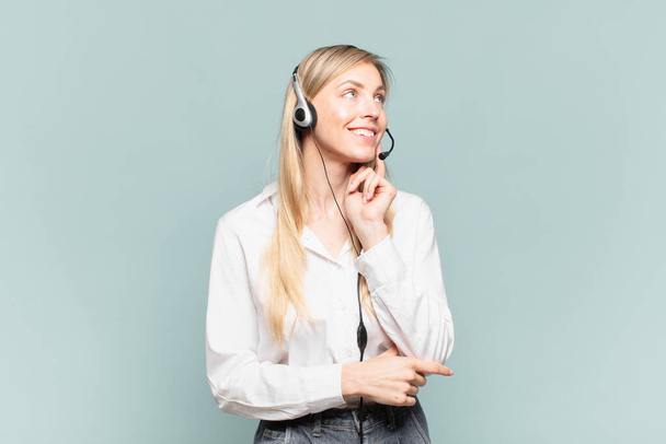 young blond telemarketer woman smiling happily and daydreaming or doubting, looking to the side - Photo, Image