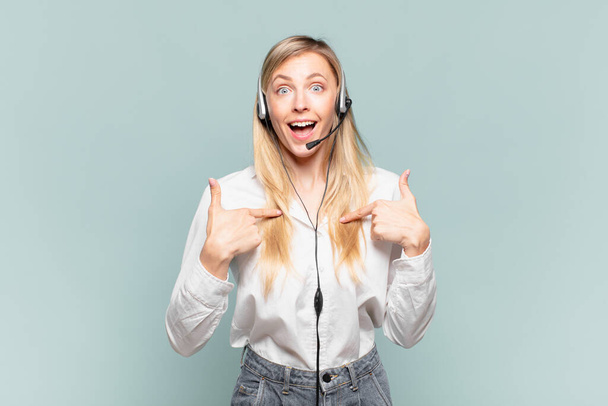 young blond telemarketer woman feeling happy, surprised and proud, pointing to self with an excited, amazed look - Foto, imagen