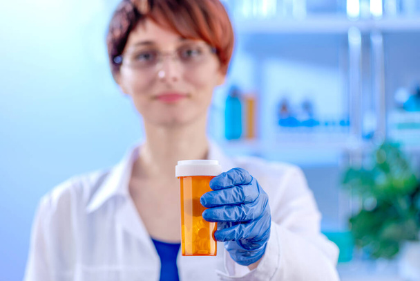 Hände in Medizinhandschuhen mit orangefarbenem Tablettenglas im Fokus, Ärztin im weißen Mantel in einem Krankenhauszimmer in Unschärfe. - Foto, Bild