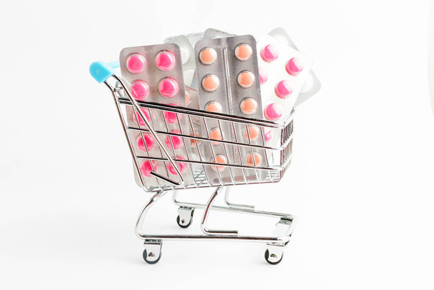 Muchas píldoras medicinales, tabletas y vitaminas en un pequeño carrito de compras del supermercado aislado sobre fondo blanco, fotografía farmacéutica tomada con enfoque suave - Foto, imagen