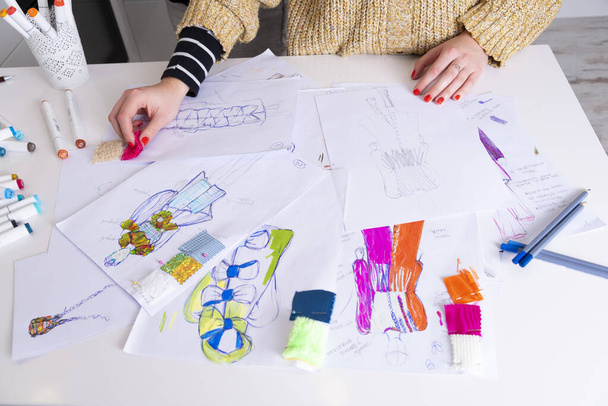 νεαρή μελαχρινή γυναίκα σχεδιάζει φορέματα μόδας σχέδιο με χρωματιστά μολύβια. Διάφορα πολύχρωμα σχέδια μόδας και σκίτσο - Φωτογραφία, εικόνα