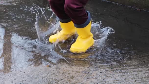 Sarı lastik çizmelerle su birikintilerinden atla. Yağmurda yürümek ve kötü havanın tadını çıkarmak çocukluğa düşmek ve ıslak su birikintilerinde koşmak. Yüzü olmayan bacak videosu. - Video, Çekim