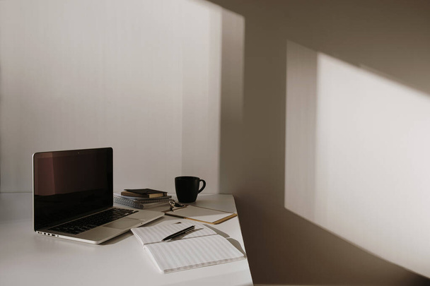 Espaço de trabalho de escritório minimalista em casa. Laptop na mesa com xícara de café, folha de papel, papelaria contra a parede branca. Trabalho, formação profissional. Sombra de luz solar na parede. - Foto, Imagem