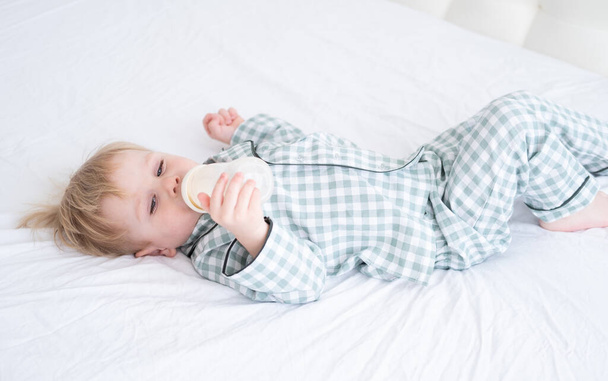 A kisgyerek pizsamában fekszik az ágyon, és üvegből issza a tejet. Őszinte otthon gyermekkori élet. - Fotó, kép
