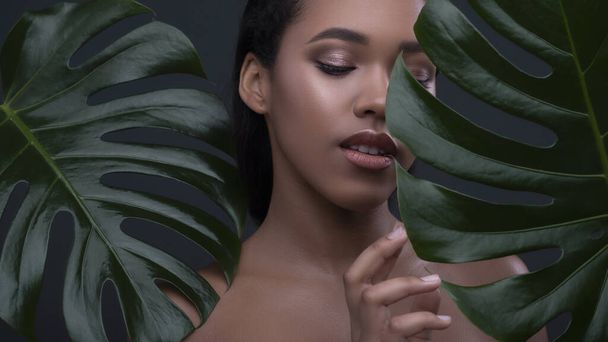 Dark skinned beautiful young woman posing sensually on a neutral green background with monstera leafs. Flawless skin with nude make up. Skin care concept portait with space for text. - Foto, Imagem