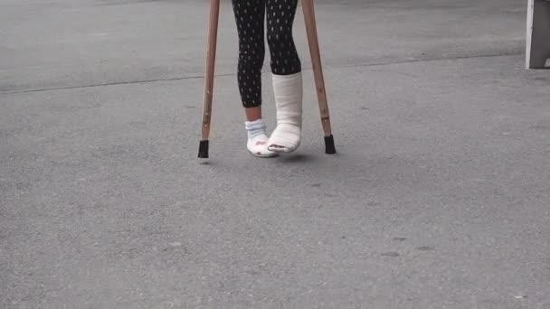 enfant blessé à la jambe, elle essaie de marcher dans la rue à l'aide de béquilles. Jambe cassée et béquilles en bois. Traumatisme, canne à pied, accident, concept de santé - Séquence, vidéo
