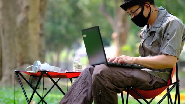 Homem em máscara usando computador portátil no fundo do jardim e relaxante nas férias - Filmagem, Vídeo