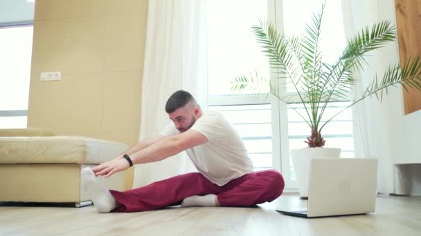 young adult bearded man doing stretching exercises at home in the living room watching video lesson or tutorial online using a laptop. Man doing fitness and sports in the morning sitting on the floor - Materiał filmowy, wideo