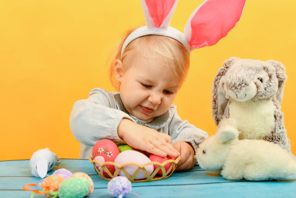 Küçük bir kız, boyalı Paskalya yumurtalarını bir sepetten çıkarıp Paskalya için oyuncak bir tavşana gösteriyor.. - Fotoğraf, Görsel