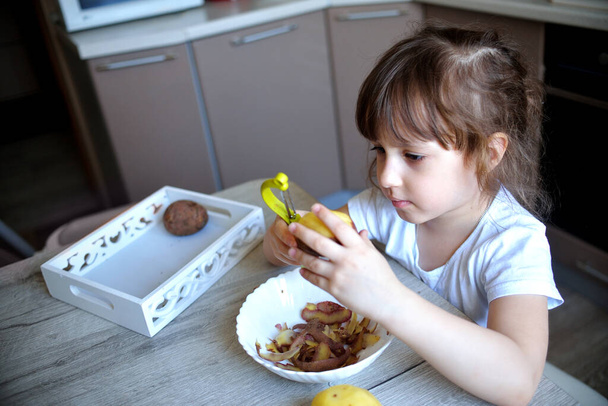 Kind schält Kartoffeln mit einem grünen Schäler. Vorschulkinder bereiten Essen zu. Montessori-Materialien für eine Lektion aus dem praktischen Leben. Lebensstil zu Hause - Foto, Bild