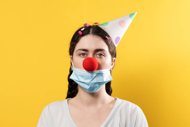April's fool day concept. Portrait of a woman in a paper cap and a false red nose with a protective mask. Yellow background. Copy space. New normal. - Photo, Image