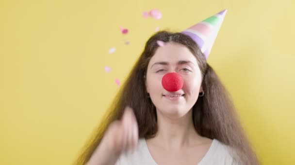 Une femme joyeuse avec un chapeau en papier et un faux nez rouge, qui vomit joyeusement des confettis. Fond jaune. Le concept du jour fou d'avril. - Séquence, vidéo