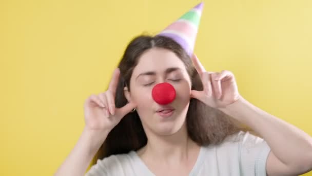 Portrait of a woman in a paper cap and a false red nose, who writhes funny and then smiles. Yellow background. April's fool day. - Footage, Video