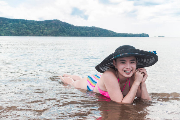 Una mujer asiática sonriente yaciendo en la arena y surfeando en aguas poco profundas en la playa. Usando un gran sombrero de sol. - Foto, imagen