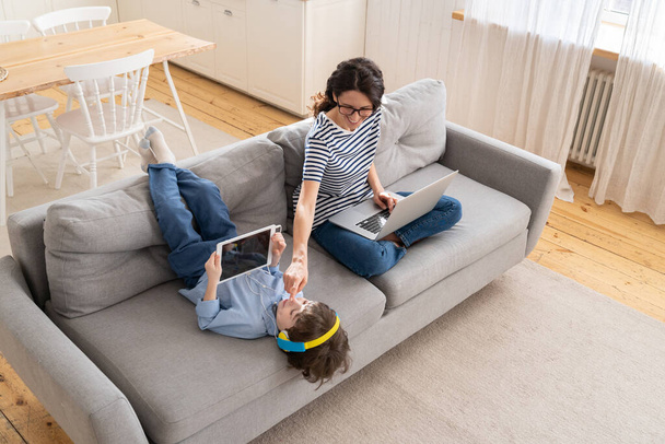 Maman freelance télétravail de bureau à domicile sur ordinateur portable assis sur canapé enfant jouer sur tablette. Verrouillage - Photo, image