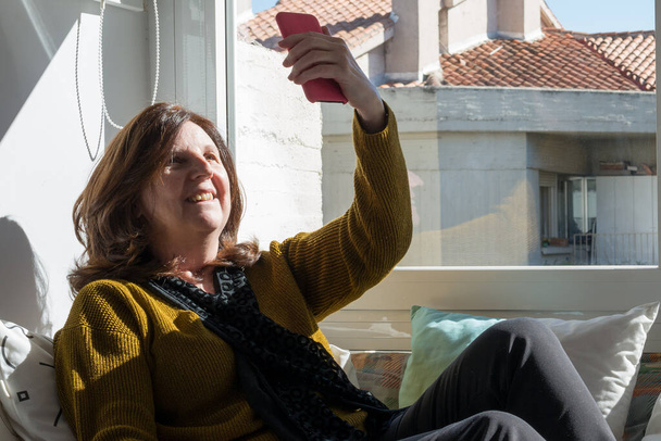 COVID中にビデオチャットのために自宅で携帯電話を使用して50歳の白人女性.窓の近くのソファに座り、太陽がガラスを通って来る. - 写真・画像