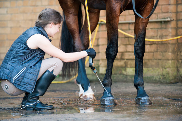 Pěkná dospívající mladá dívka na koni mytí kopyt a nohy koně ve sprše po tréninku. Pulzující pestrobarevné léto venku horizontální obrázek. - Fotografie, Obrázek