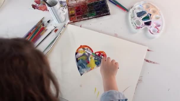 Klein kind tekent kleurrijk hart op wit papier. Kunstacademie concept. Bovenaanzicht van een meisje kind zitten aan de tafel en schilderen met aquarelverf verf. 4k-beelden - Video